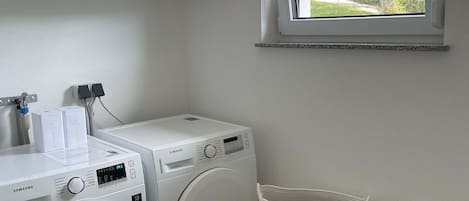 A laundry room with two white Samsung washing machines, a window with a view of greenery, and two woven baskets.