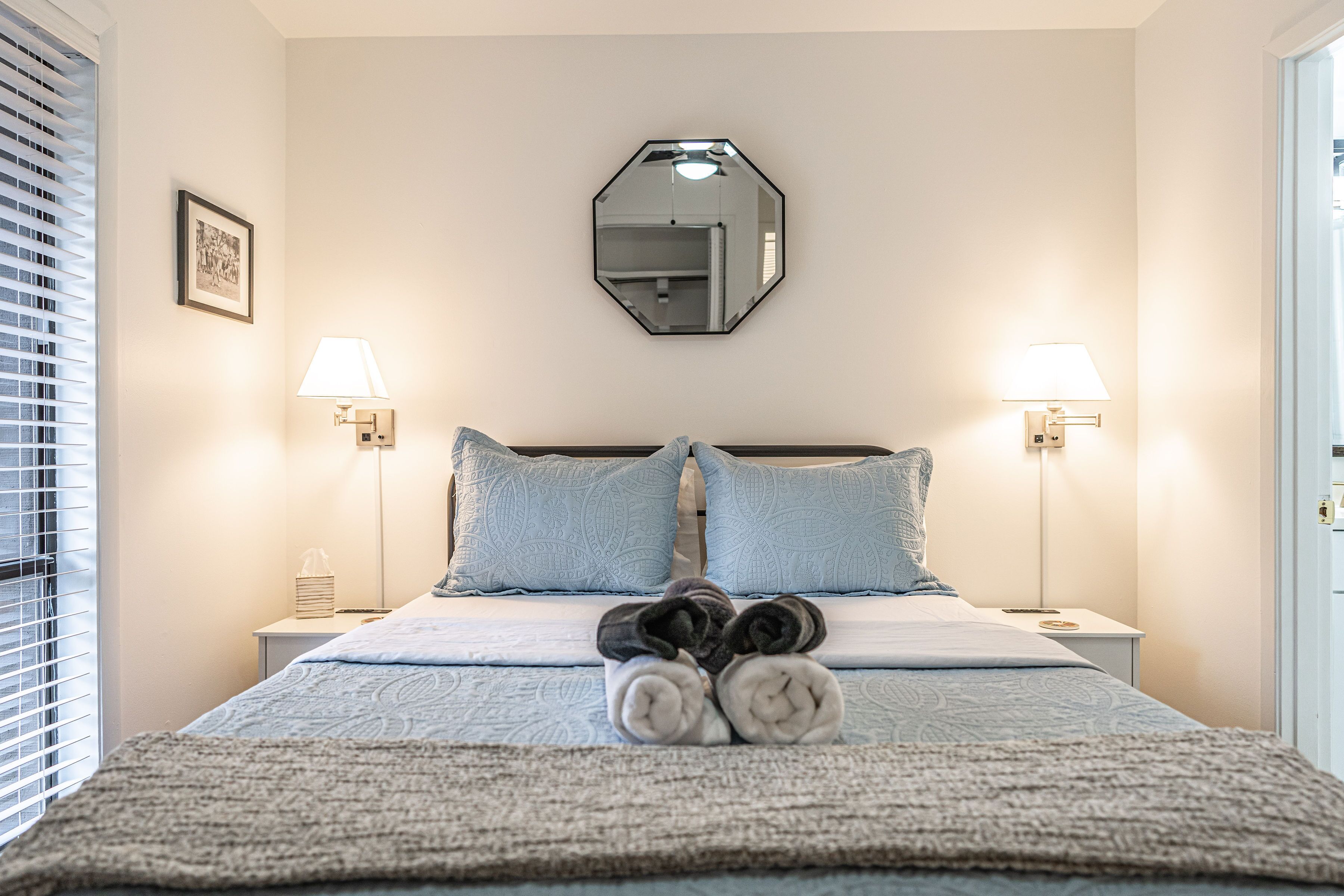 A neatly made bed with rolled towels, two bedside lamps, a framed picture, and a mirror on the wall.