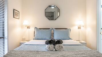 A neatly made bed with rolled towels, two bedside lamps, a framed picture, and a mirror on the wall.