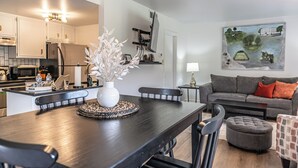 A modern kitchen with white cabinets, a dark wooden dining table with a vase of white flowers, and a grey sofa in the living area. - Long Drive On No. 5 - 1BR Condo w/ GREAT Golf Views, Minutes From Clubhouse (Pinehurst)