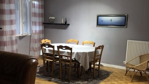 Uma sala de jantar com uma mesa posta para quatro pessoas, cadeiras de madeira, um tapete estampado, um sofá de couro marrom, uma televisão na parede e uma janela com cortinas.