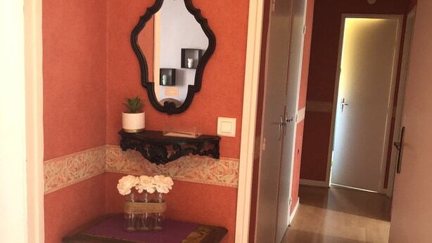 Um corredor com chão de madeira, uma pequena mesa com um vaso de flores, um espelho fixado na parede e uma porta ao fundo.