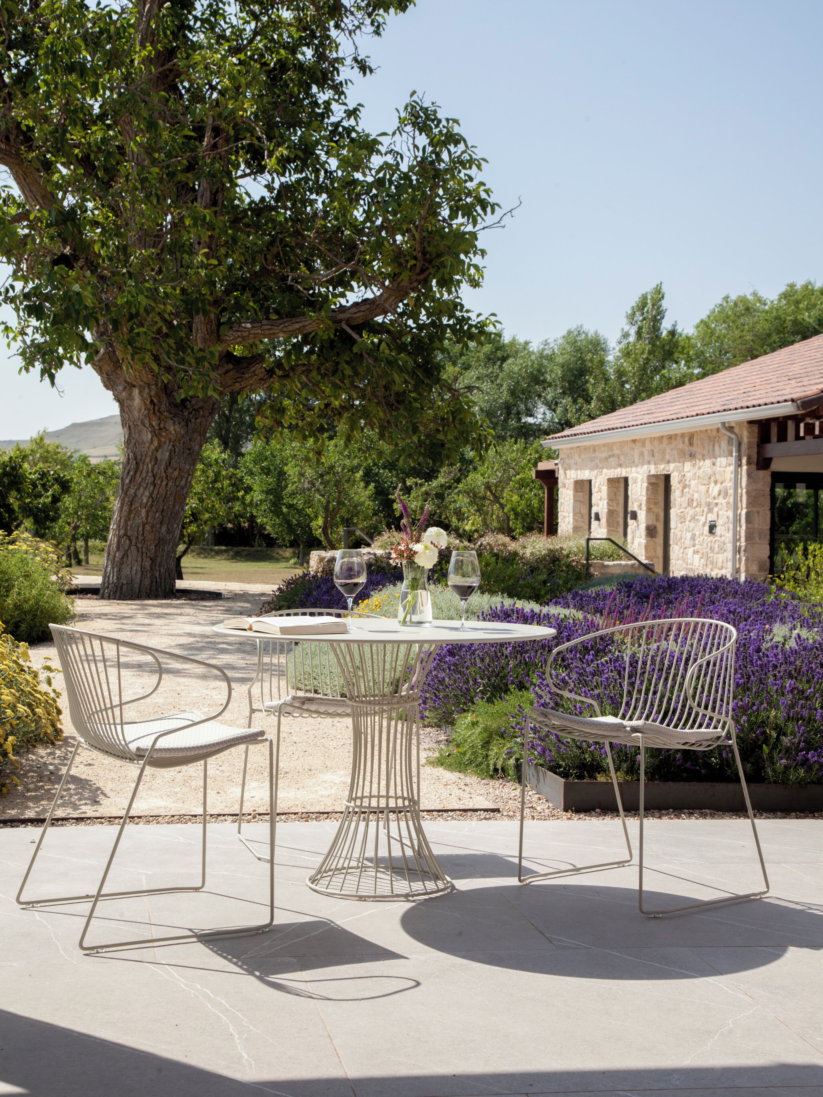 An outdoor patio with a table and chairs, surrounded by a garden with purple flowers and a large tree.