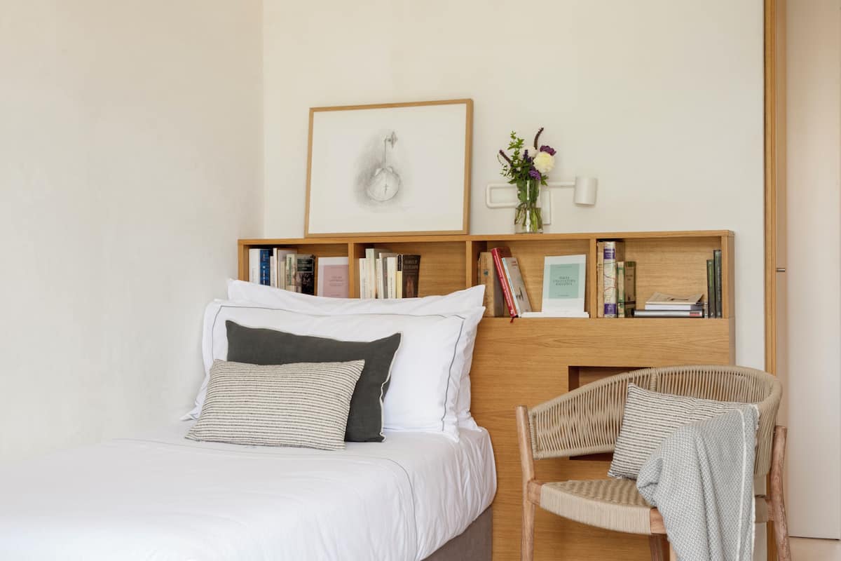 A modern bedroom with a bed, a wooden nightstand with books, a framed picture, and a wicker chair.