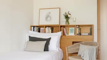 A modern bedroom with a bed, a wooden nightstand with books, a framed picture, and a wicker chair.