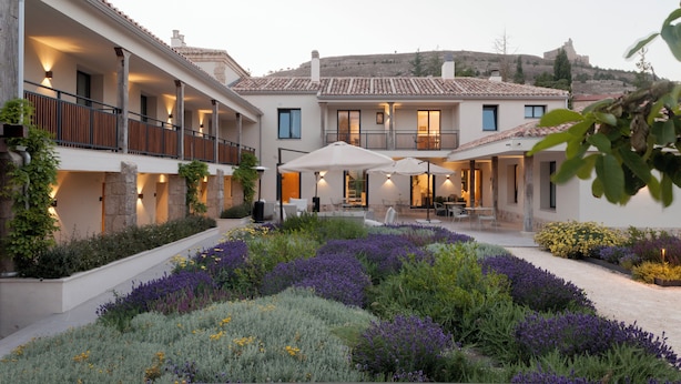 Un patio con piscina, rodeado de un jardín con flores moradas y un edificio de techo con tejas.