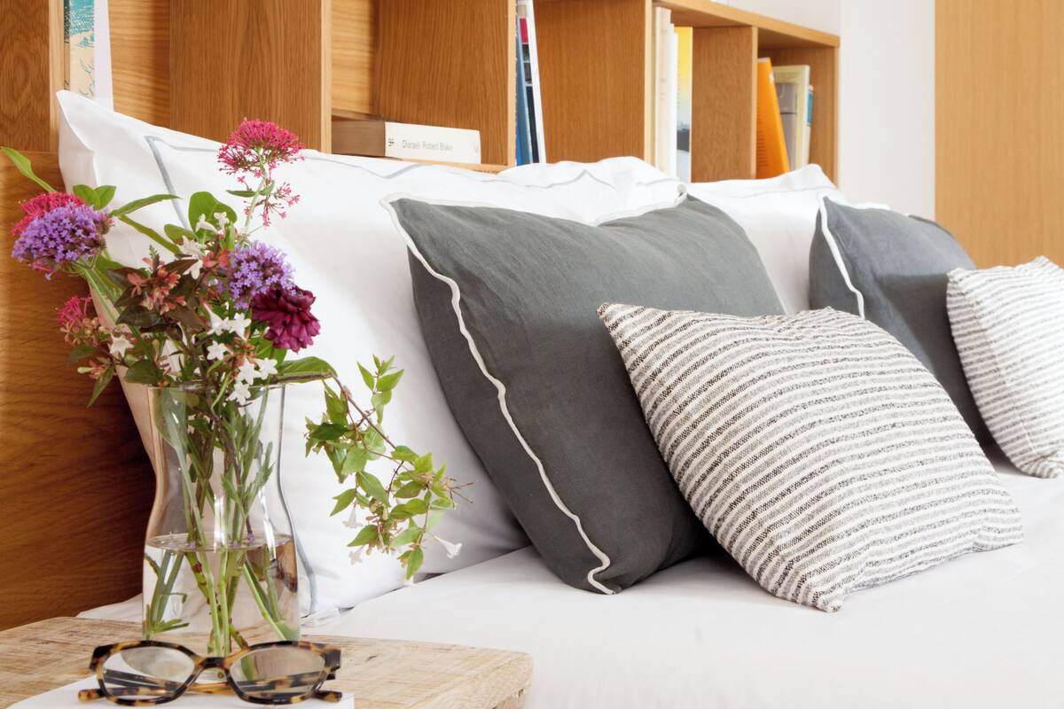 A neatly arranged bedroom with a bed, pillows, a bedside table with glasses and a book, a wooden shelf with books and a vase of flowers.