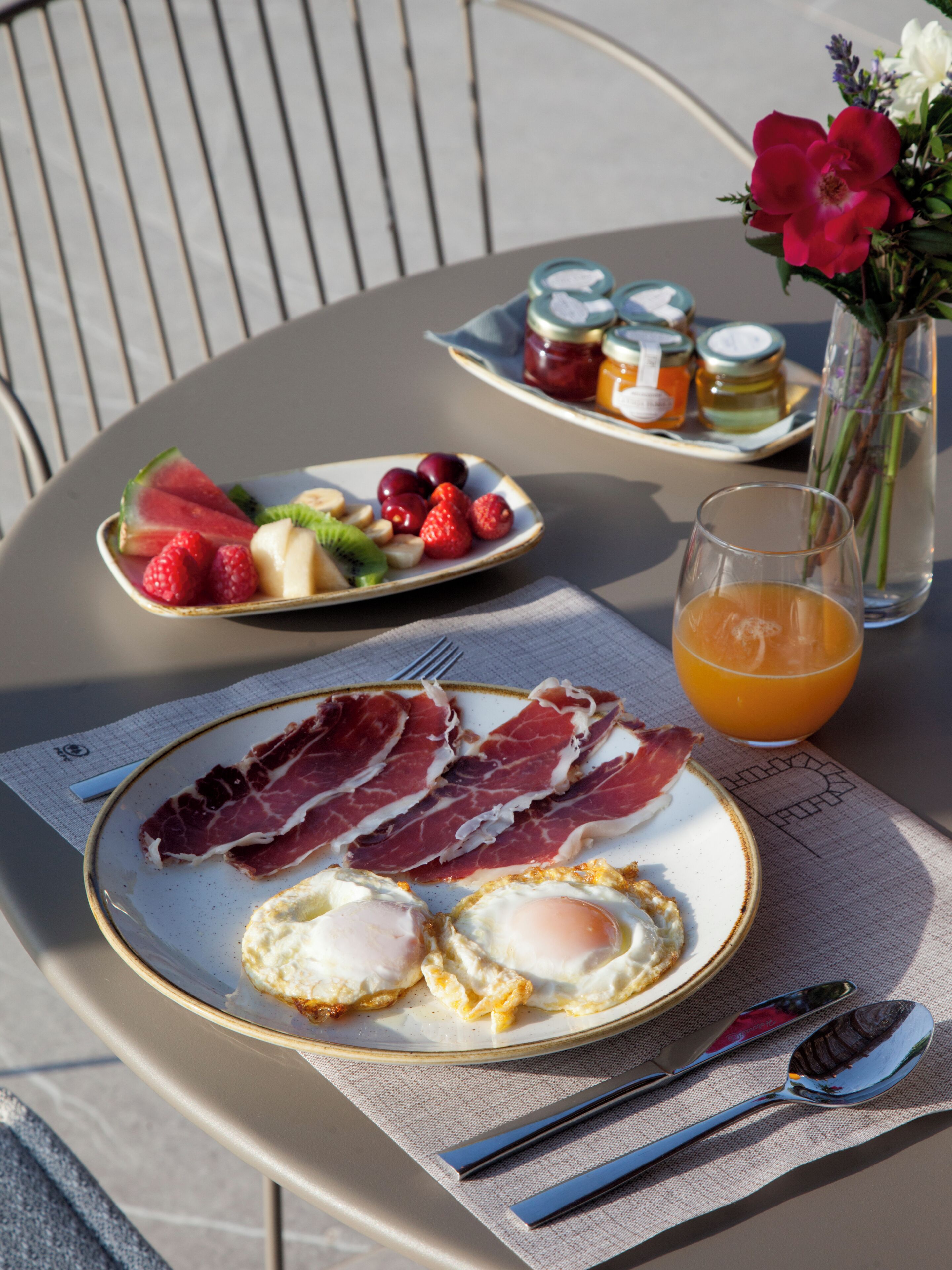 En frukostuppdukning med tallrikar fyllda med ägg, skinka och frukt, ett glas apelsinjuice och en vas med blommor på ett bord.