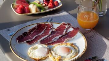 En frukostuppdukning med tallrikar fyllda med ägg, skinka och frukt, ett glas apelsinjuice och en vas med blommor på ett bord.