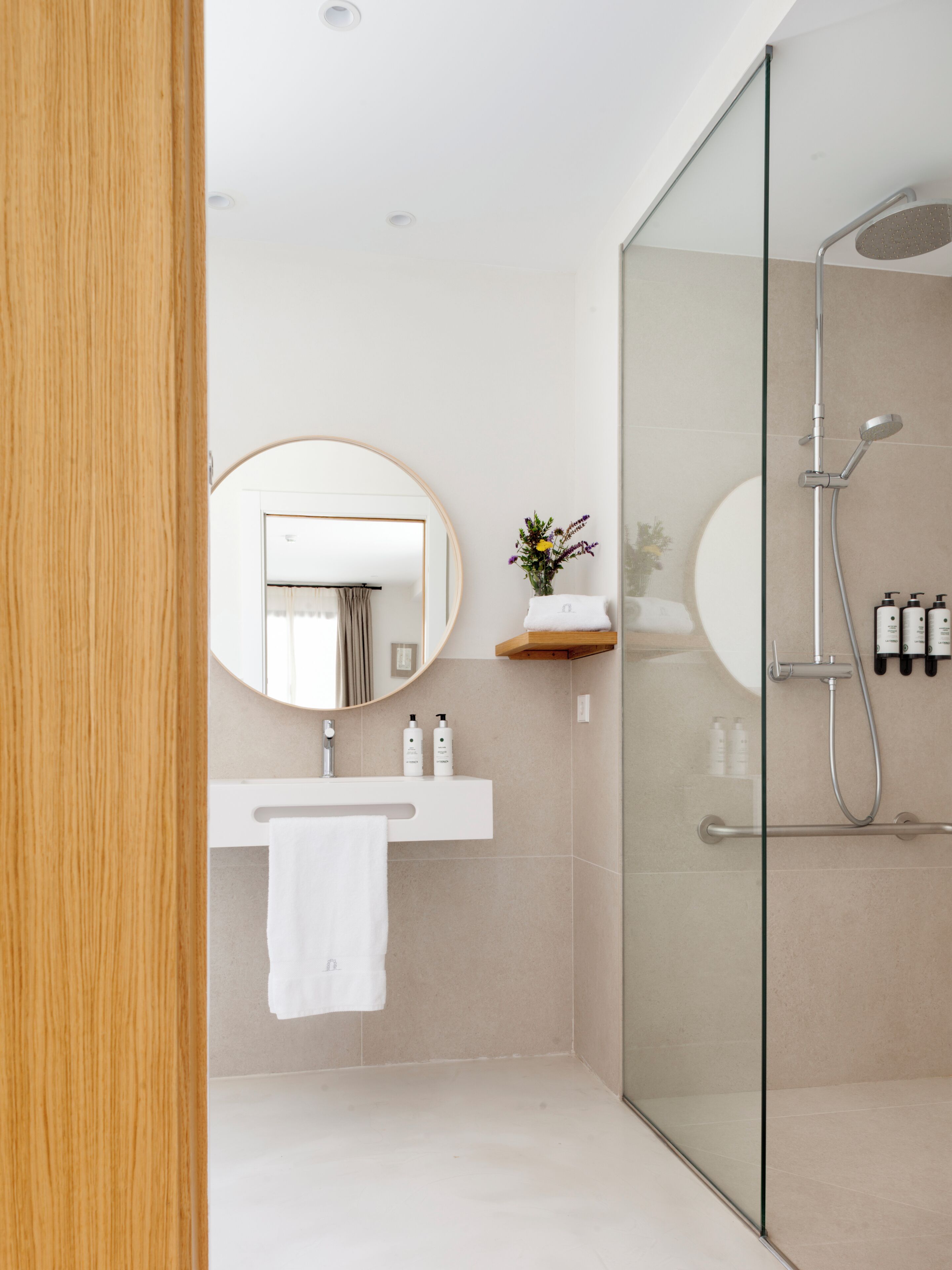 A modern bathroom with a glass shower enclosure, a round mirror, a white sink with a towel, and a small shelf with toiletries.