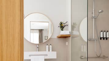 A modern bathroom with a glass shower enclosure, a round mirror, a white sink with a towel, and a small shelf with toiletries.