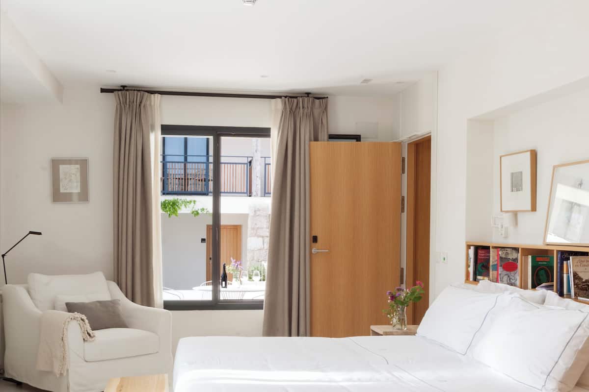 A modern bedroom with a large bed, a sofa, a wooden side table, and a view of the outside balcony.