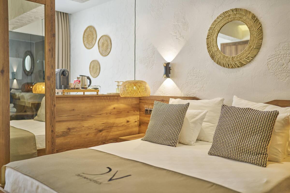 Quarto de hotel com cabeceira de madeira, cama com roupa de cama branca, almofadas estampadas, espelho redondo e pratos decorativos fixados na parede.