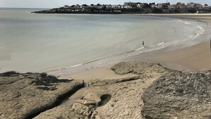 A coastal beach with rocky foreground, calm water, and a town in the background.