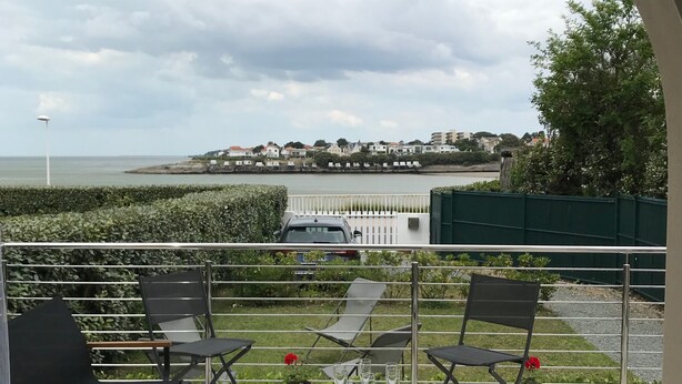 An outdoor patio with a table set for two, two lounge chairs, and a view of the sea and buildings in the distance.