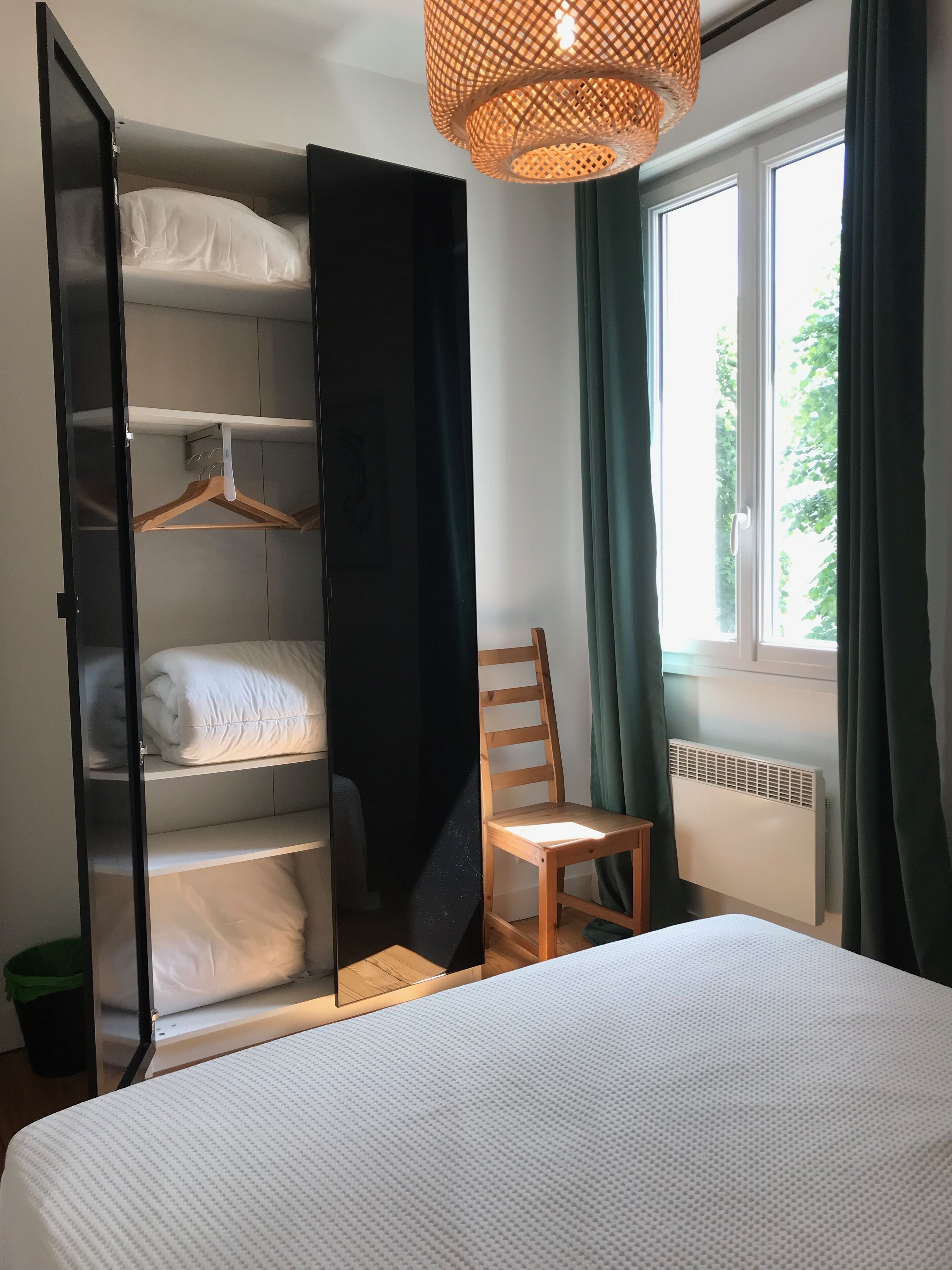 Une chambre à coucher comprenant un lit, une armoire avec plusieurs étagères, une chaise, un luminaire suspendu et une fenêtre avec des rideaux.
