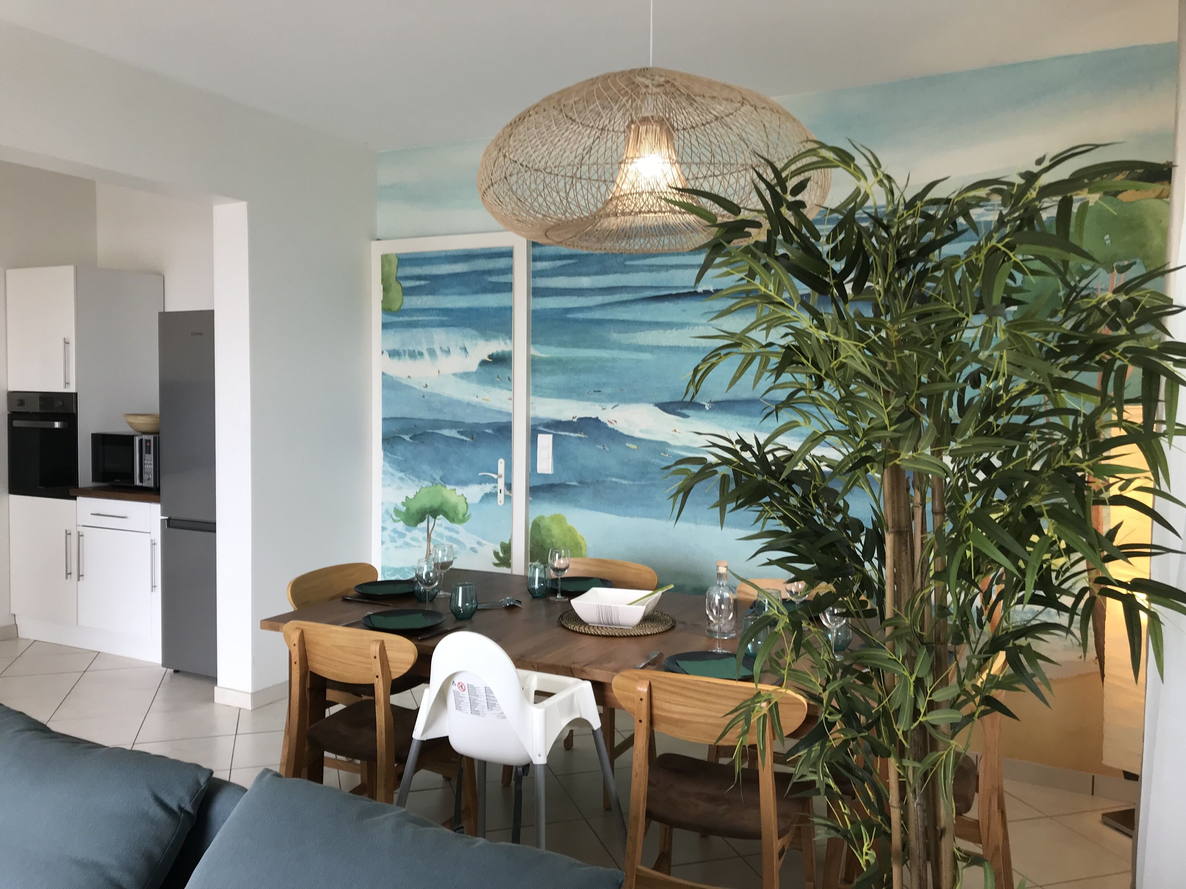 Un coin repas avec une table dressée pour quatre personnes, entourée de chaises, d’une grande plante en pot et d’une fresque murale représentant un paysage marin.