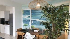 A dining area with a table set for four, surrounded by chairs, a large potted plant, and a wall mural depicting a seascape.