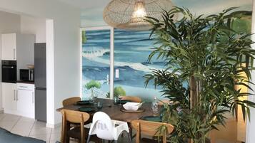 Un coin repas avec une table dressée pour quatre personnes, entourée de chaises, d’une grande plante en pot et d’une fresque murale représentant un paysage marin.