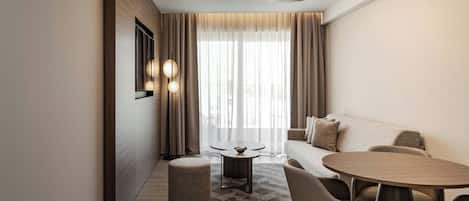 A modern living room with a round wooden table, two chairs, a sofa, and a small round ottoman. There is a large window with sheer curtains and a ceiling light fixture.