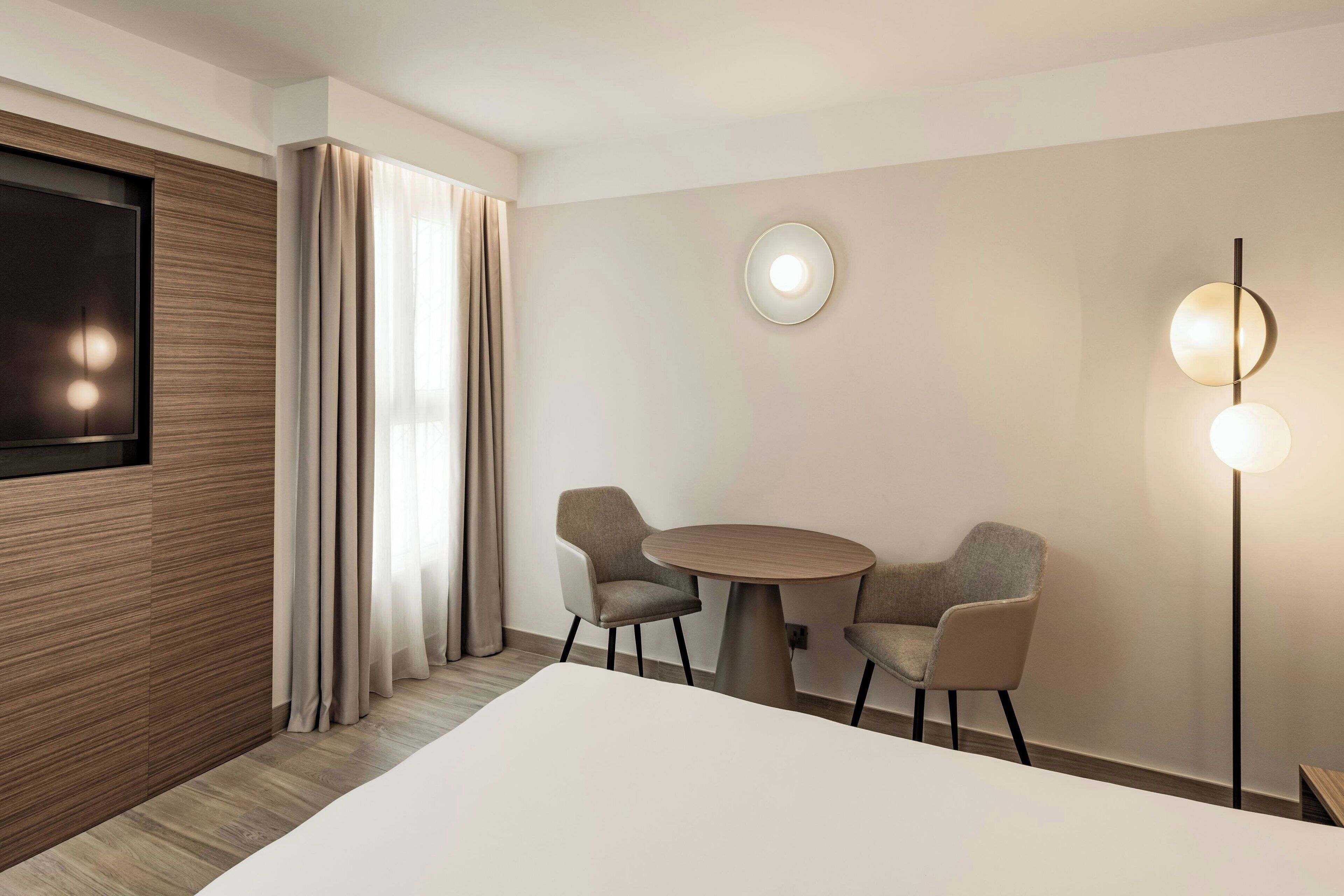 A hotel room with a large bed, a TV mounted on a wooden panel, a round table with two chairs, a floor lamp, and a wall-mounted light fixture.