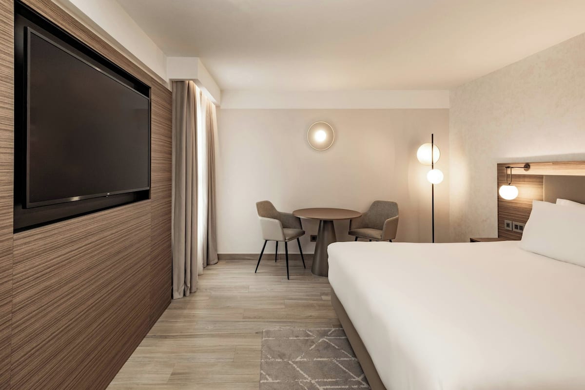 A modern hotel room with a large bed, a flat-screen TV mounted on the wall, a round dining table with chairs, and a bedside table with a lamp.