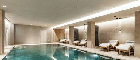 A modern indoor swimming pool with clear blue tiles, lounge chairs, and recessed lighting.