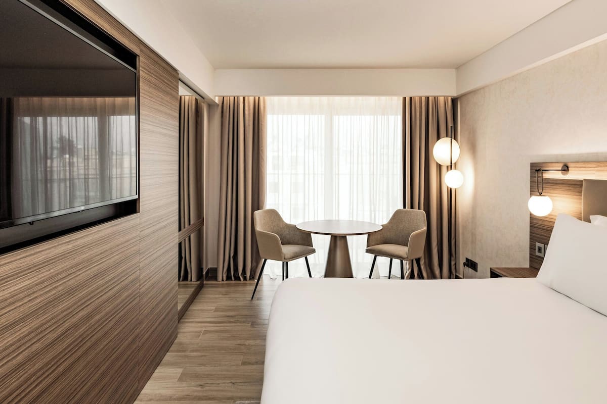 A hotel room with a large bed, a TV mounted on the wall, a round table with two chairs, and a window with curtains.