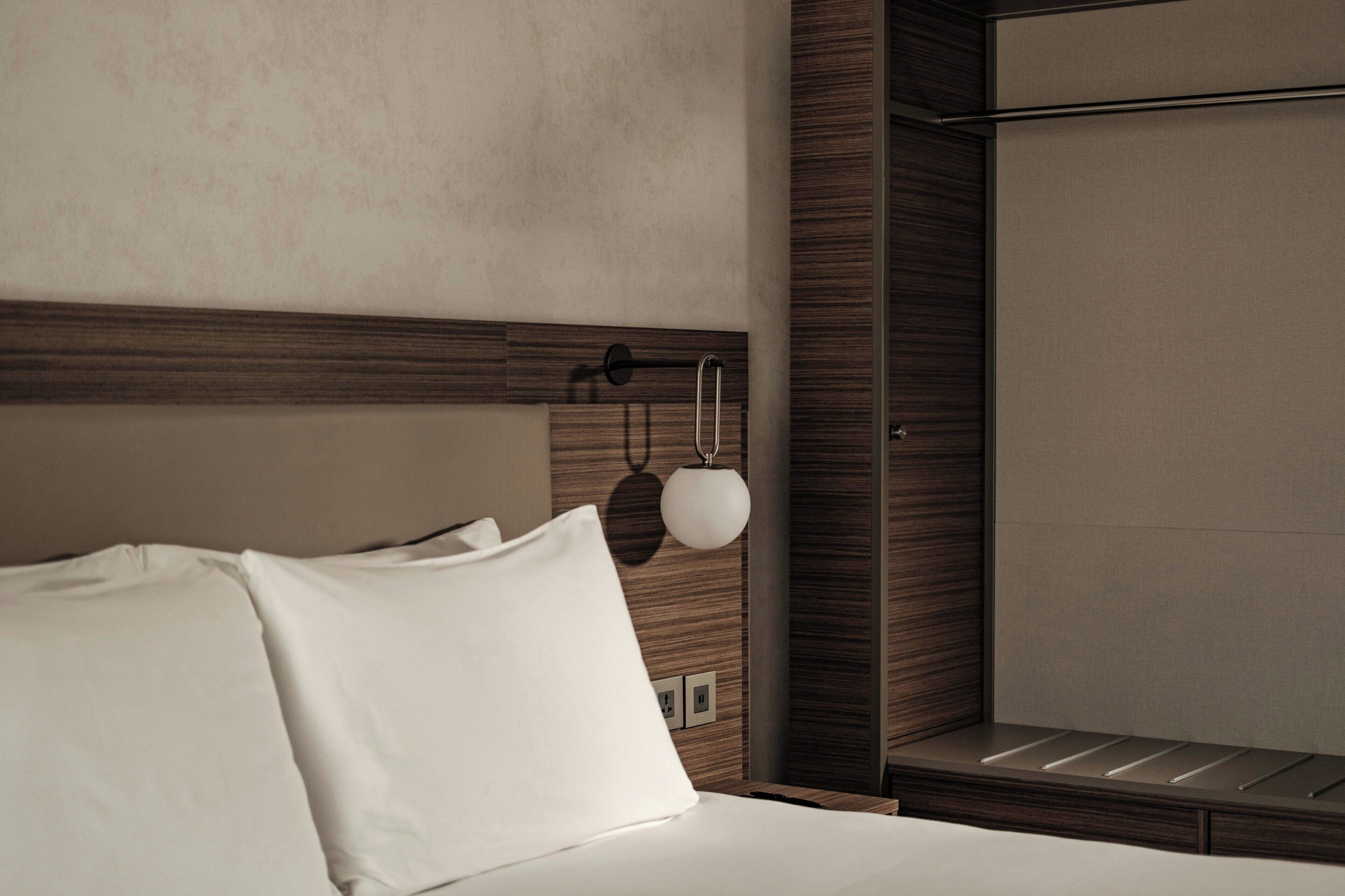 Habitación de hotel con una cama bien hecha, cabecera de madera, lámpara de pared y un ventanal grande.