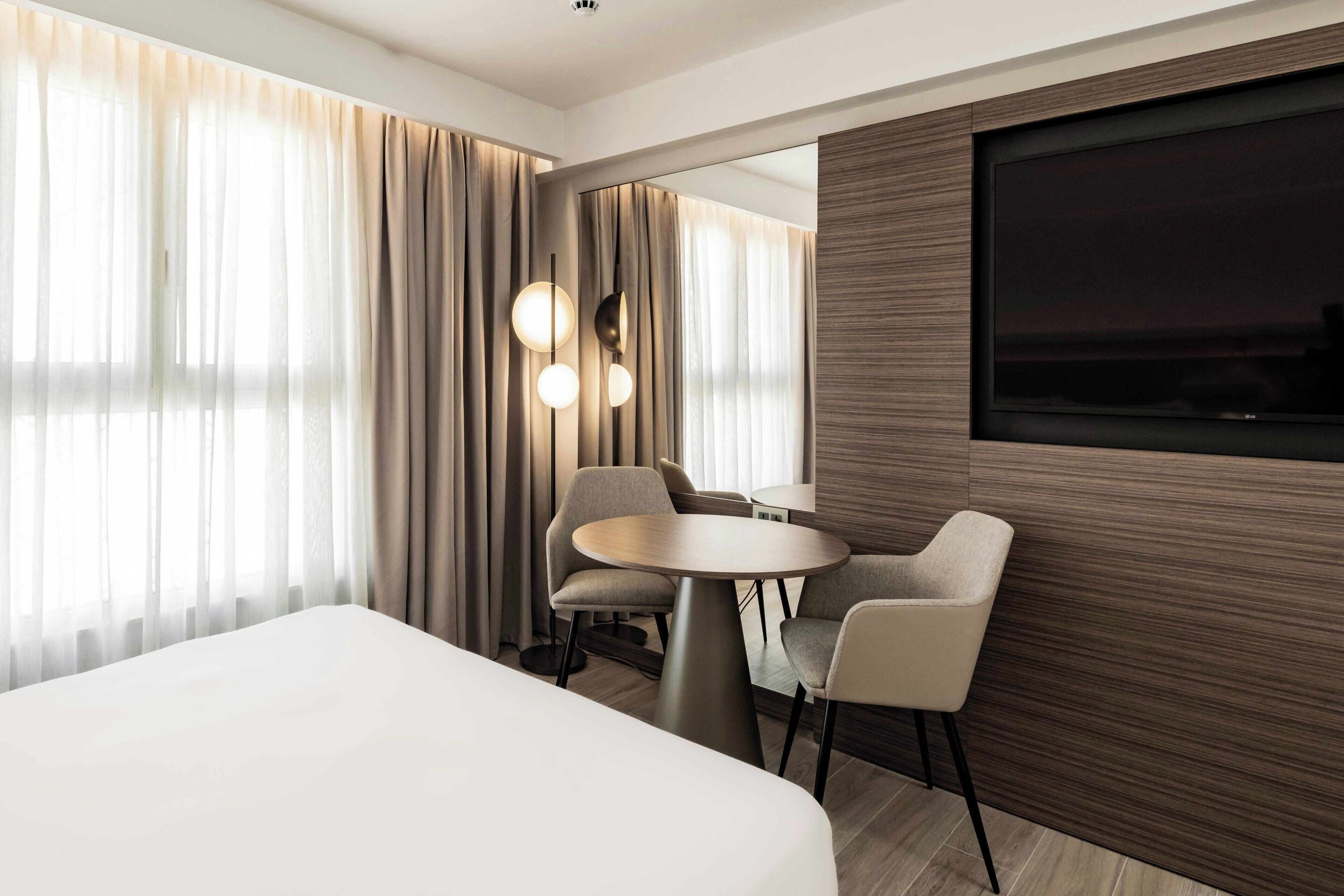 A modern hotel room with a large bed, a round dining table with chairs, a TV mounted on the wall, and a large window with curtains.