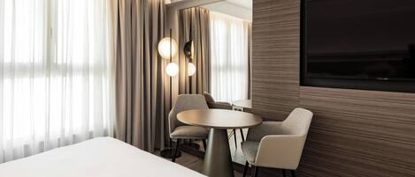 A modern hotel room with a large bed, a round dining table with chairs, a TV mounted on the wall, and a large window with curtains.