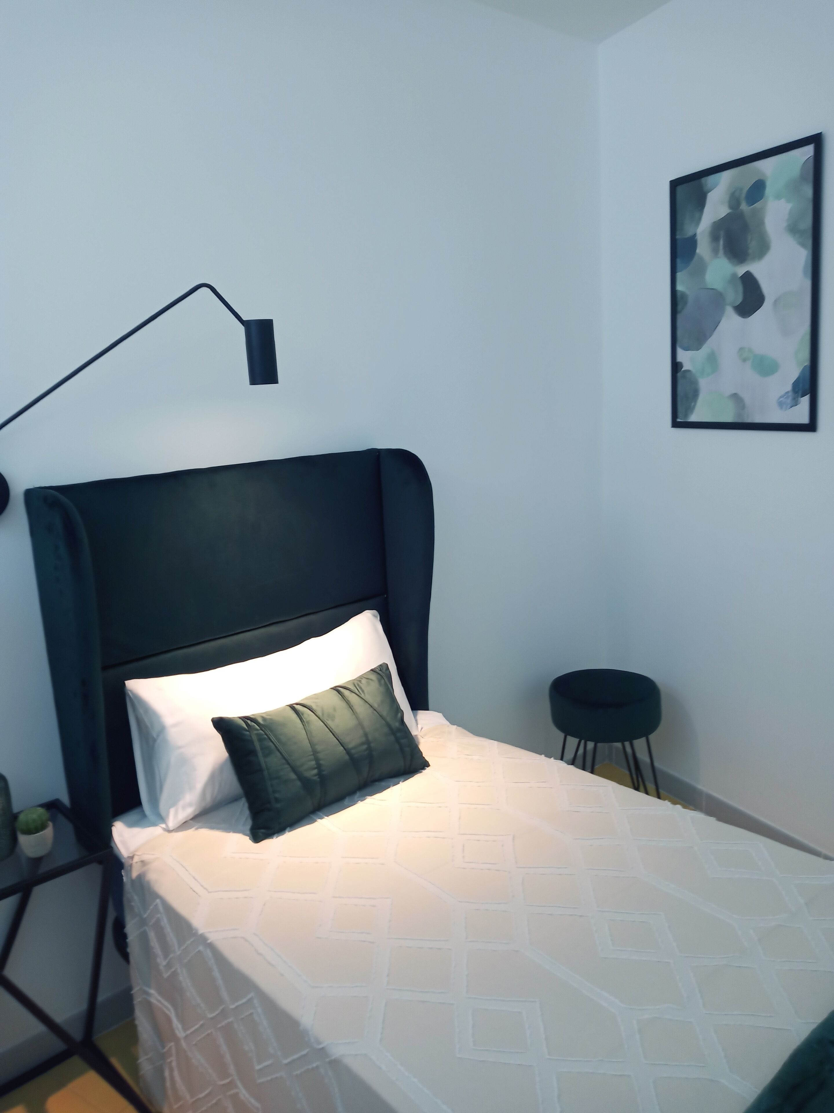 A neatly made bed with a white and patterned bedspread, a dark headboard, and a single green pillow. A black floor lamp is positioned to the left of the bed.