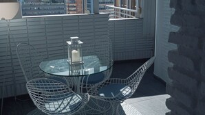 A balcony with a glass table and wire chairs, overlooking a cityscape with buildings and a mountain.
