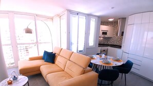 A modern living room with a sofa, a dining table, and chairs. There is a kitchen area with cabinets and a microwave.