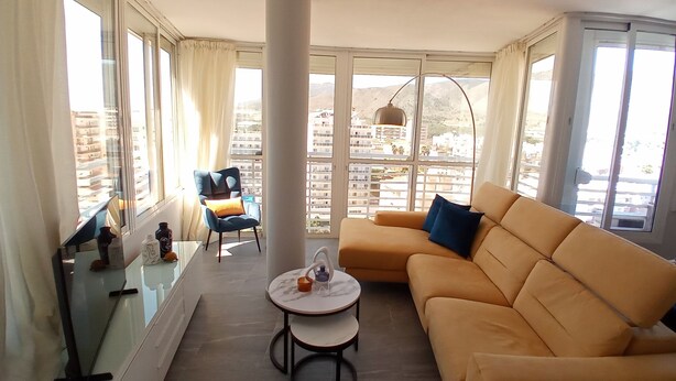 A living room with a sofa, a chair, a small table, and a view of buildings through large windows.