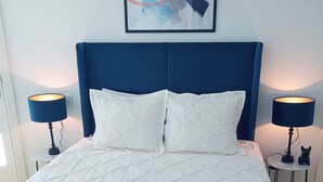 A neatly made bed with a white quilted duvet, two white pillows, and a blue throw blanket. There are two bedside lamps with black shades, and a framed abstract painting above the bed.