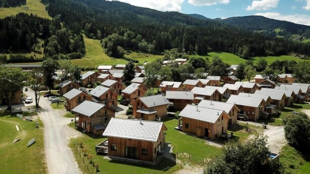 Un gruppo di casette in legno con tetti spioventi, disposte in modo compatto con una strada sterrata in primo piano e colline boschive sullo sfondo.