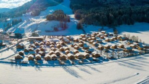 Un villaggio innevato con case di legno disposte a cerchio, circondato da montagne e un cielo sereno.