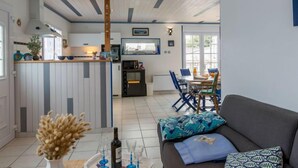 Uma cozinha moderna com uma ilha central, uma área de jantar com mesa e cadeiras, e uma sala de estar com sofá e almofadas.
