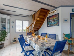 Un comedor con escalera de madera, una mesa dispuesta con platos, tazas y una botella, rodeada de sillas azules.
