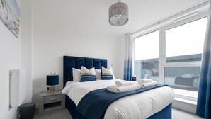 A modern bedroom with a large bed, a bedside table with a lamp, a window with curtains, and a view of the outside. - Stylish Penthouse- Guests Favourite. Just Amazing! (London)