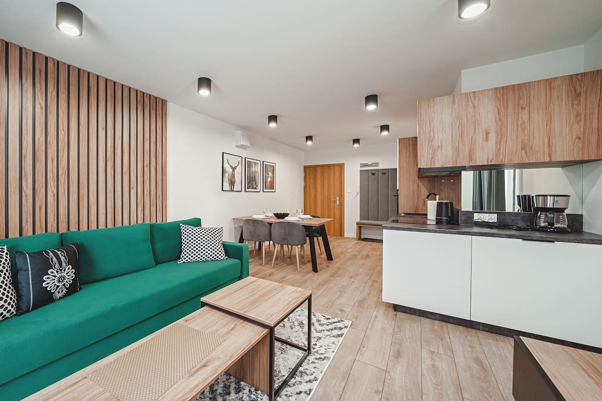 A modern living room with a green sofa, a dining table, and a kitchen area with wooden cabinets and a coffee machine.