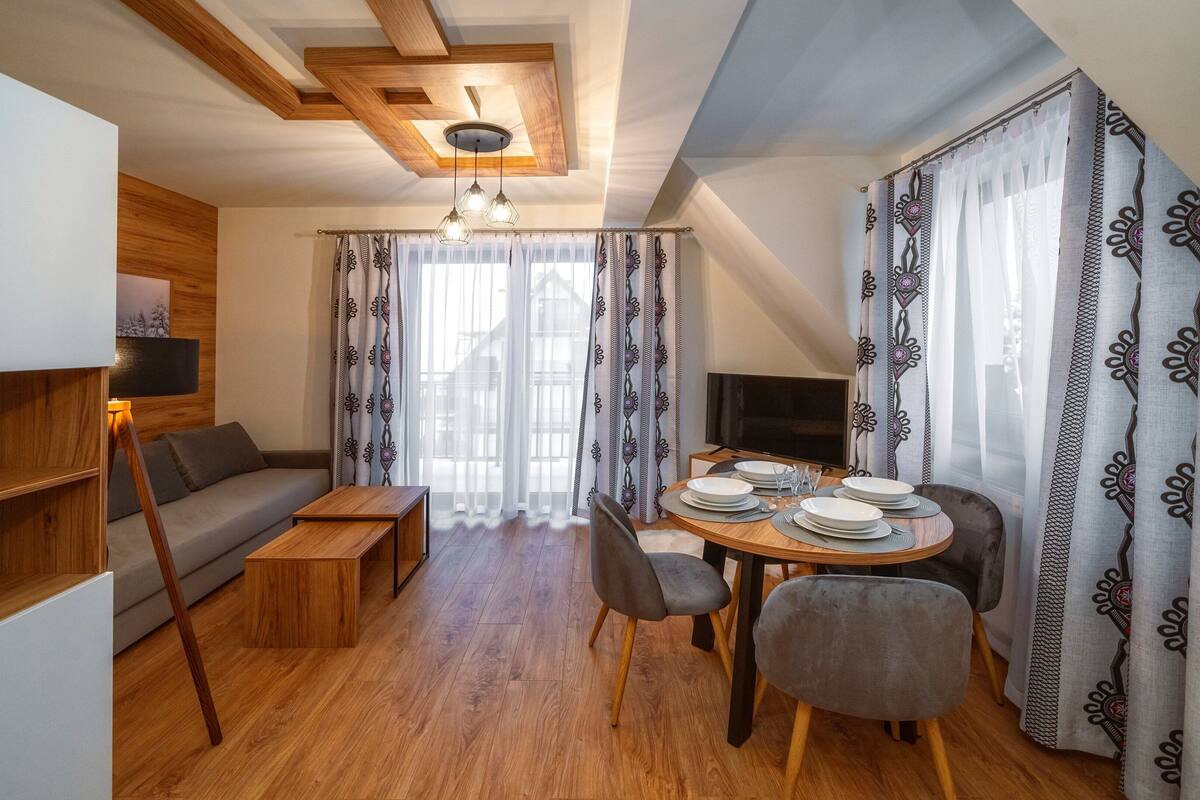 A modern living room with a wooden ceiling, a sofa, a dining table set for two, and patterned curtains.