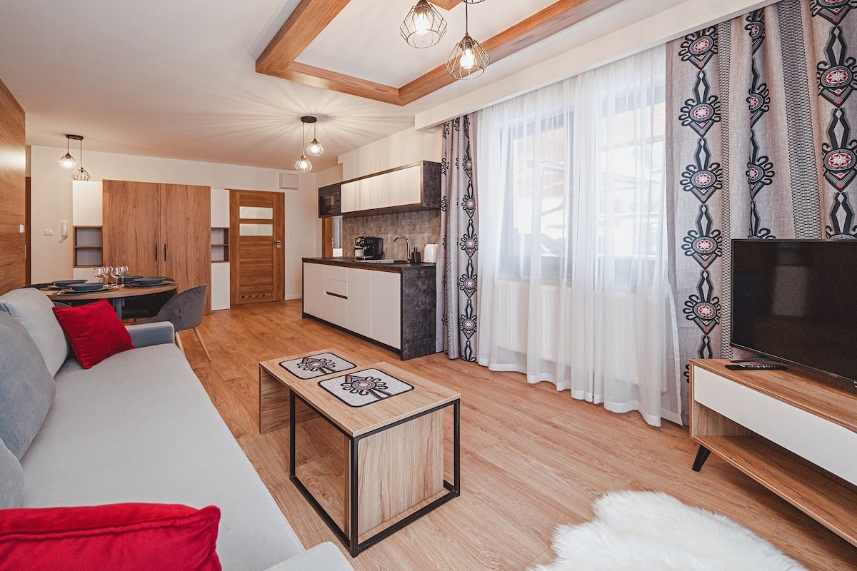 A modern living room with a sofa, a coffee table, and a TV. There is a kitchen area with a microwave and a dining table with chairs.