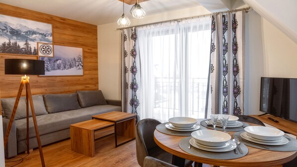 Un salon moderne avec un plancher en bois, un canapé gris, une table basse en bois et un coin repas avec une table ronde dressée pour quatre personnes.