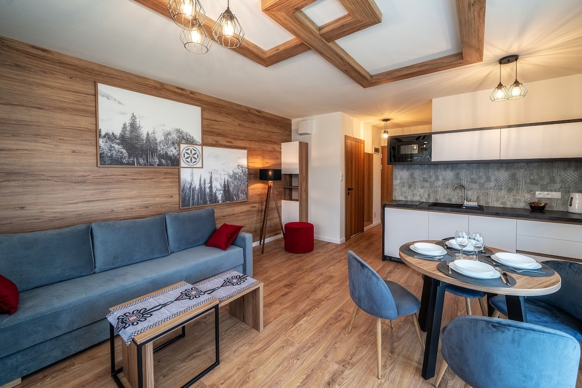 A modern living room with a sofa, dining table, and chairs, a kitchen area with cabinets and a countertop, and a wooden ceiling with skylights.