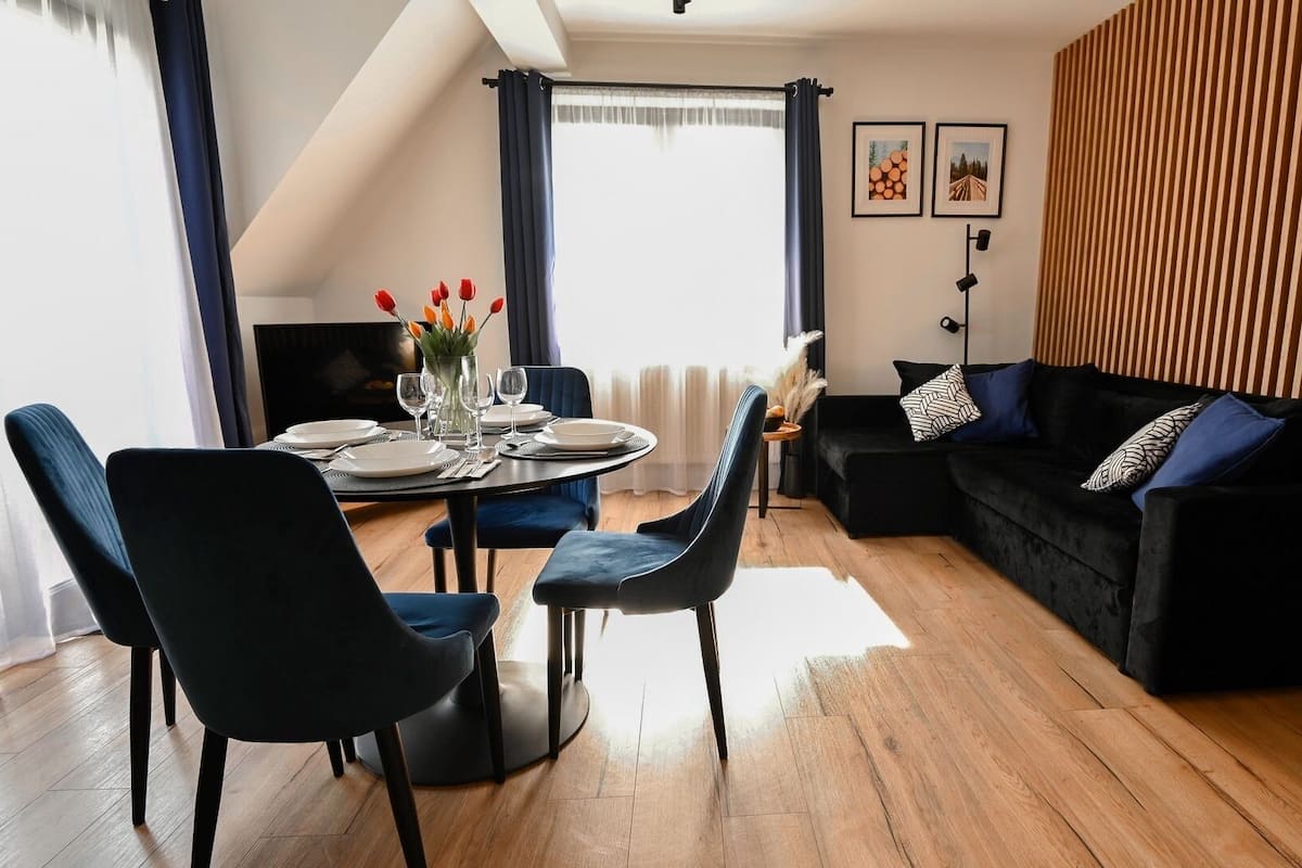 A modern living room with a dining area, featuring a black sofa, blue chairs, a round dining table set for four, and a wooden slatted wall.