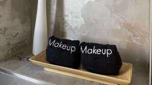 Un baño con pared texturizada, un jarrón blanco con una planta verde y dos toallas negras con el bordado 'Makeup' sobre una bandeja de madera.