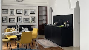 A modern living room with a blue sofa, yellow chairs, a dining table set for four, and a kitchen with a black island. - Ortigia Suite 1939 (Siracusa)
