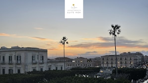 A cityscape at sunset with classical architecture, palm trees, and a clear sky. - Ortigia Suite 1939 (Siracusa)